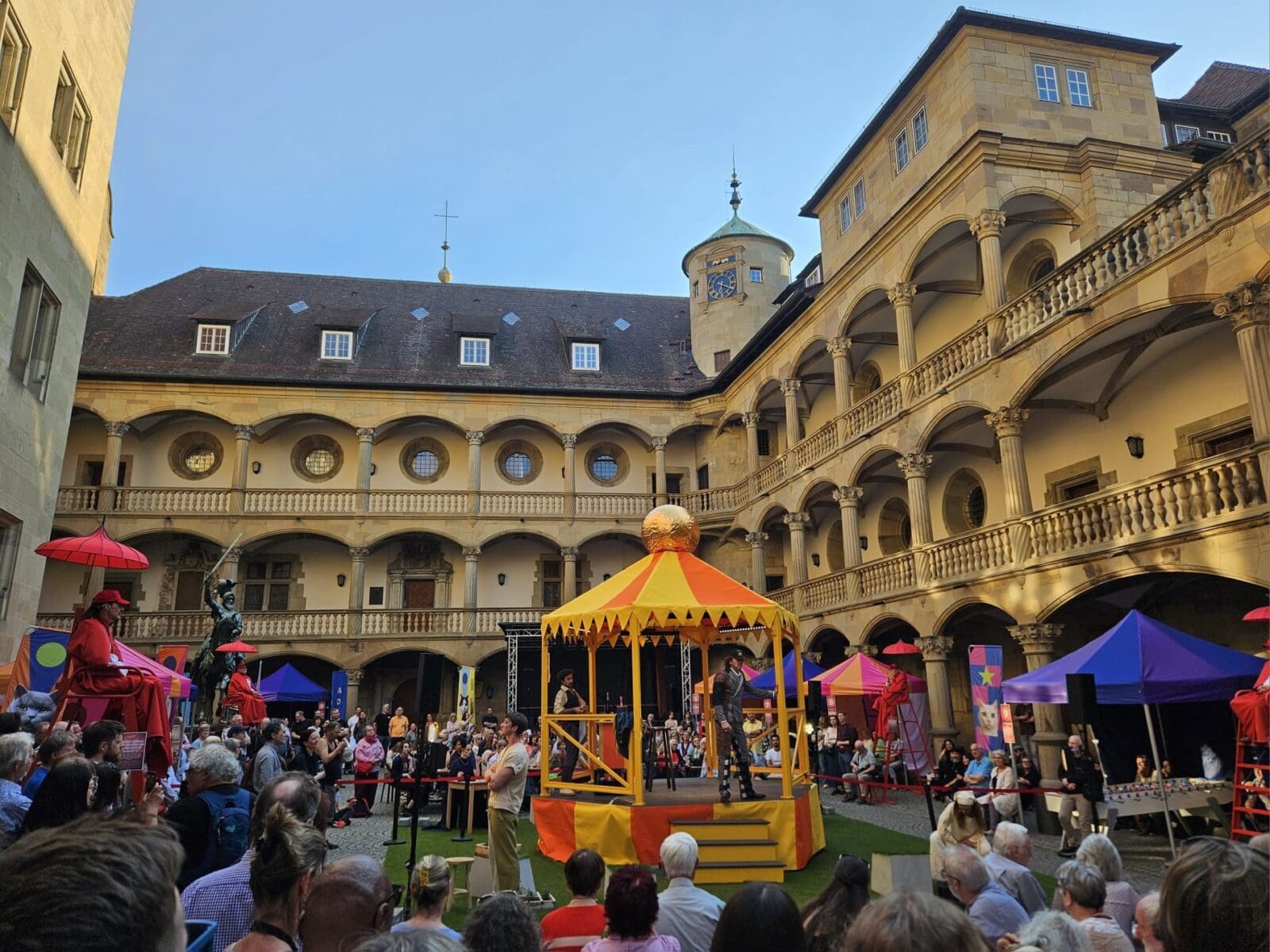 Eine runde Bühne steht im Hof des Alten Schlosses, davor Publikum, das eine Spielszene beobachtet.
