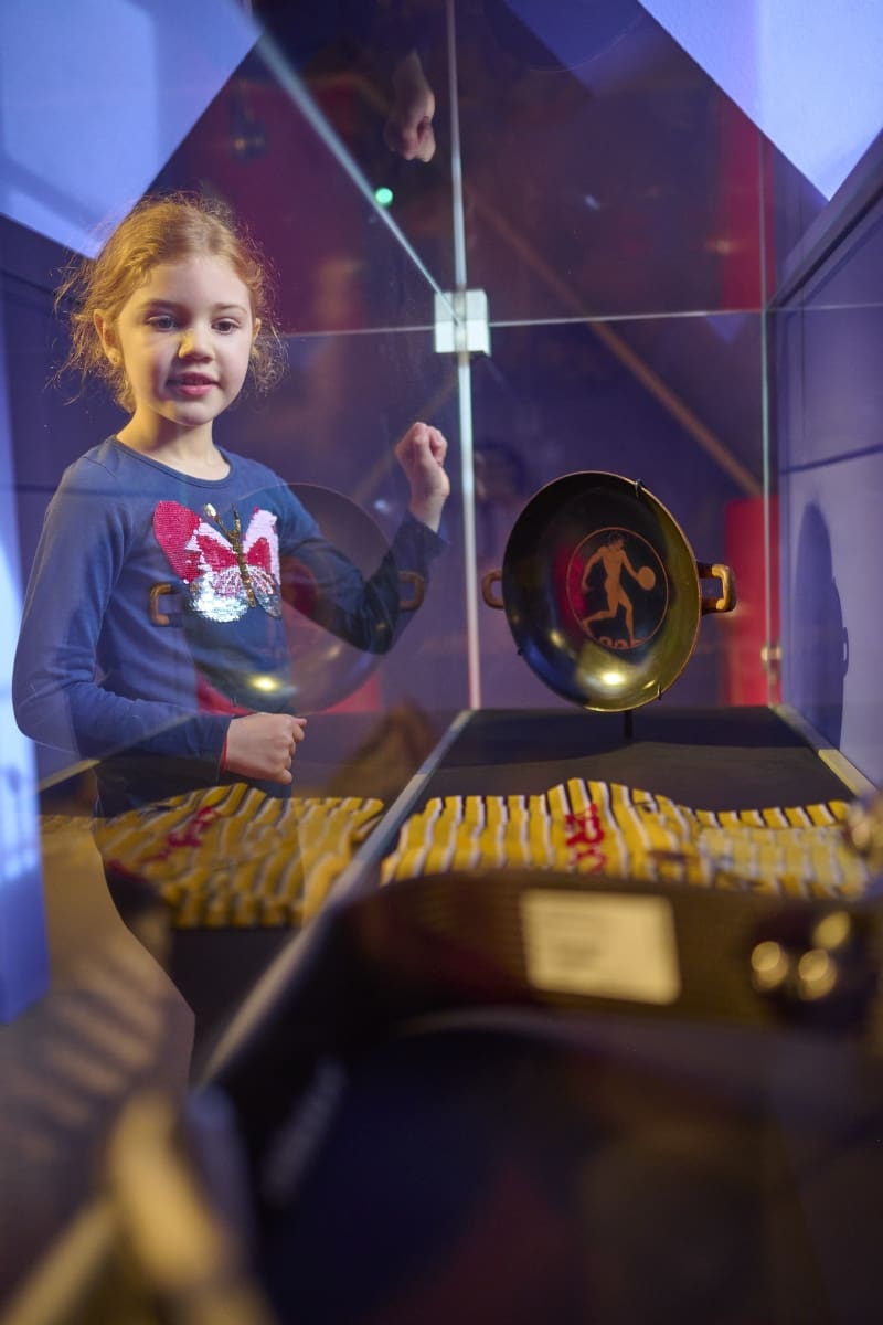 Eine Vitrine zum Thema Sportlicher Wettstreit zeigt eine antike Trinkschale mit Diskuswerfer sowie Trikot und Sportprothese des Paralympics-Weltrekord-Trägers im Weitsprung, Léon Schäfer.