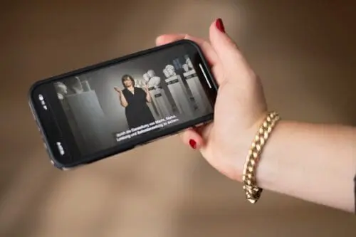 Eine Hand hält ein Smartphone. Darauf ist ein Ausschnitt der Mulitmediaguide mit Tour in Deutscher Gebärdensprache durch die Schausammlung "Wahre Schätze - Antike" zu sehen.