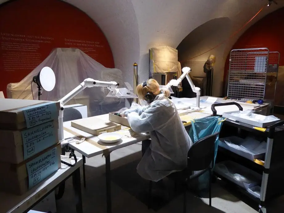 Eine Person sitzt an einem Tisch und packt römisches Fundmaterial in neue, säurefreie Kartons um.