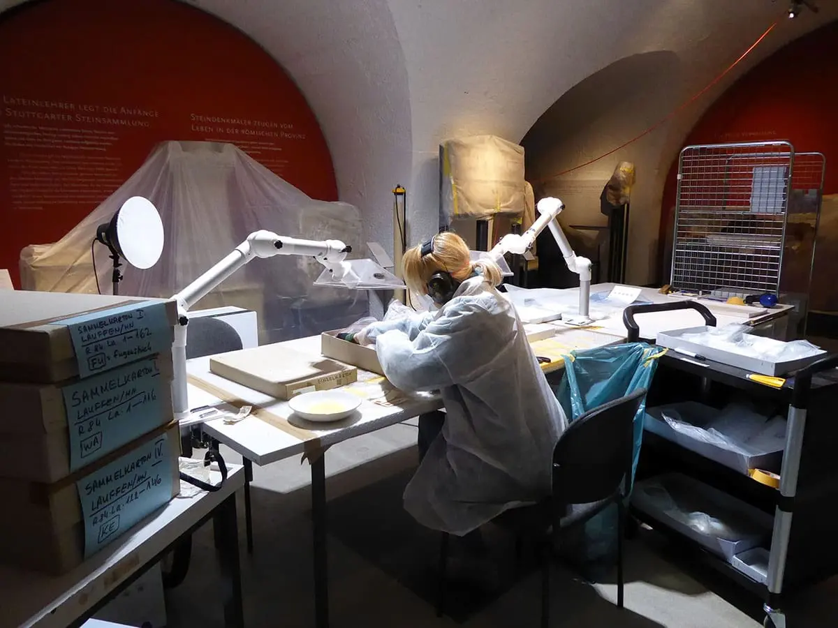 Eine Person sitzt an einem Tisch und packt römisches Fundmaterial in neue, säurefreie Kartons um.