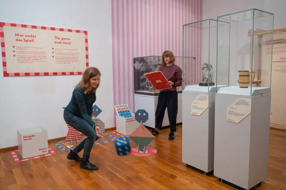 Zwei Frauen in einer Ausstellung „We Are Family“ mit übergroßem Würfel und Spielkarte