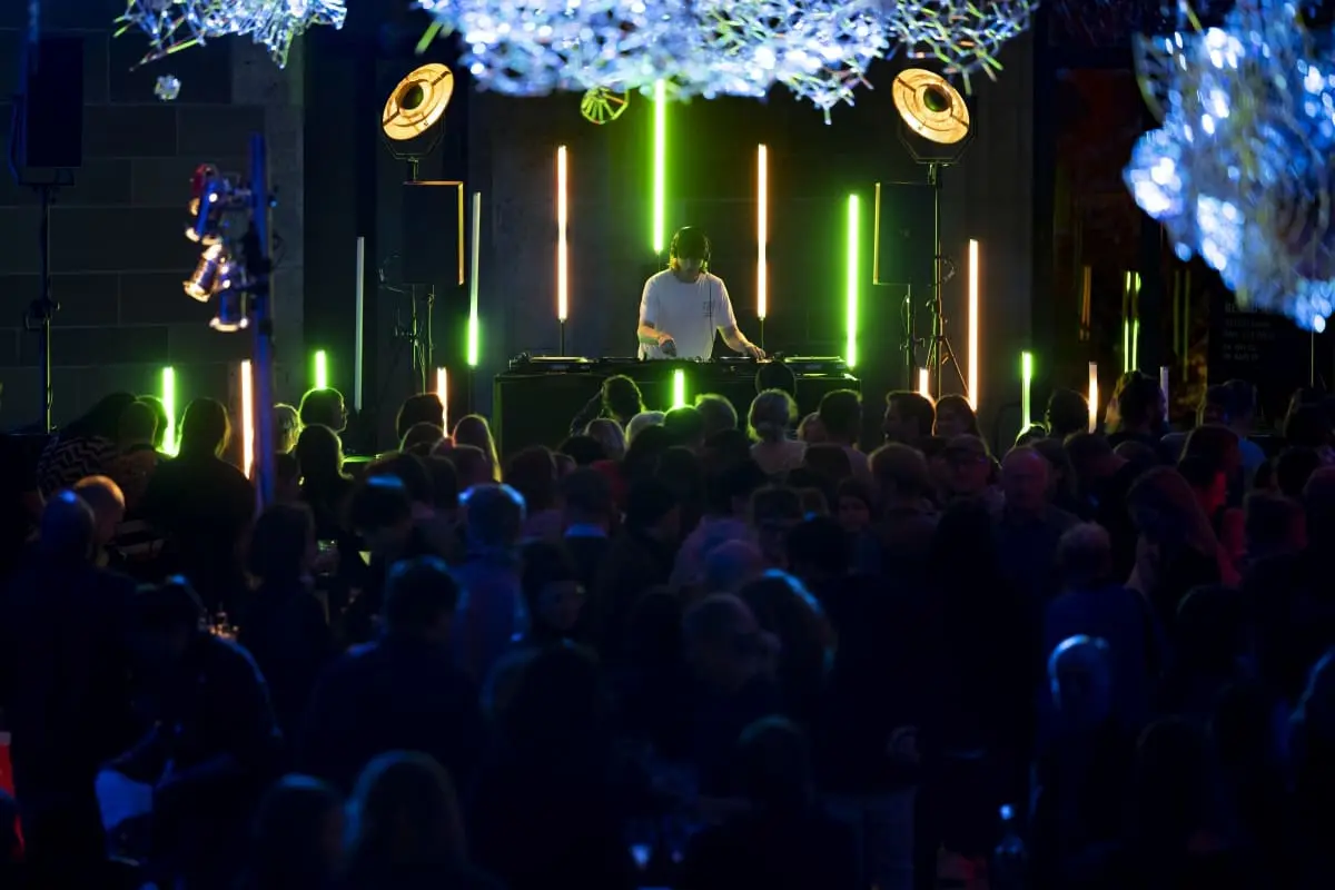 Auf der Fotografie ist ein eine Menschenmenge vor einem DJ zu sehen, der im Museumsfoyer Dürnitz auflegt. Orangene, grüne und blaue Lichter rahmen ihn, während die tanzende Menschenmenge im dunklen verschwimmt.