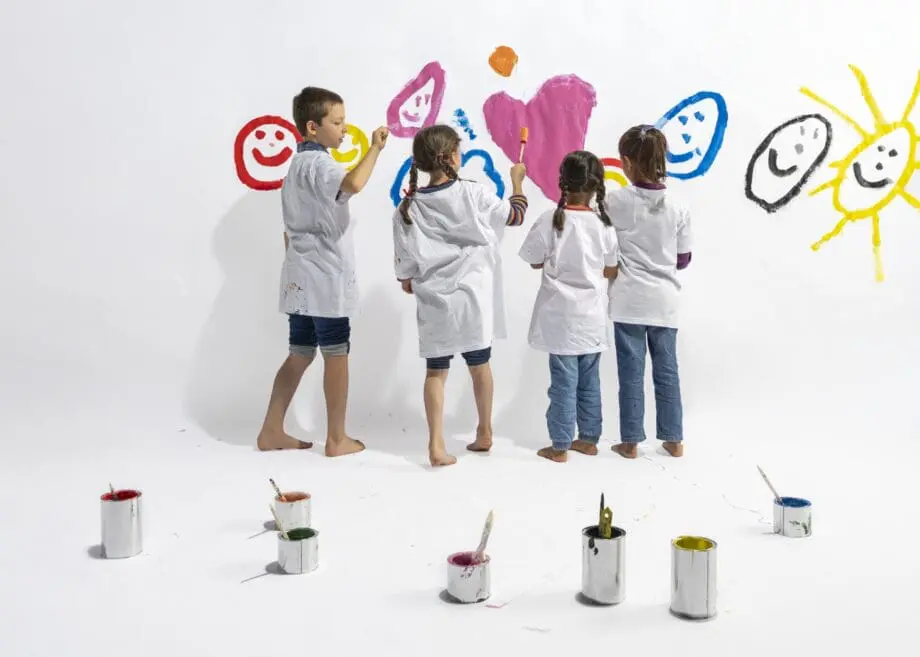 Vier Kinder, ein Junge und drei Mädchen, malen mit bunter Farbe Herzen und Smileys an eine weiße Wand. Die Aufnahme zeigt die Kinder von der Rückenansicht und sie tragen weiße Oberteile und sind barfuß.